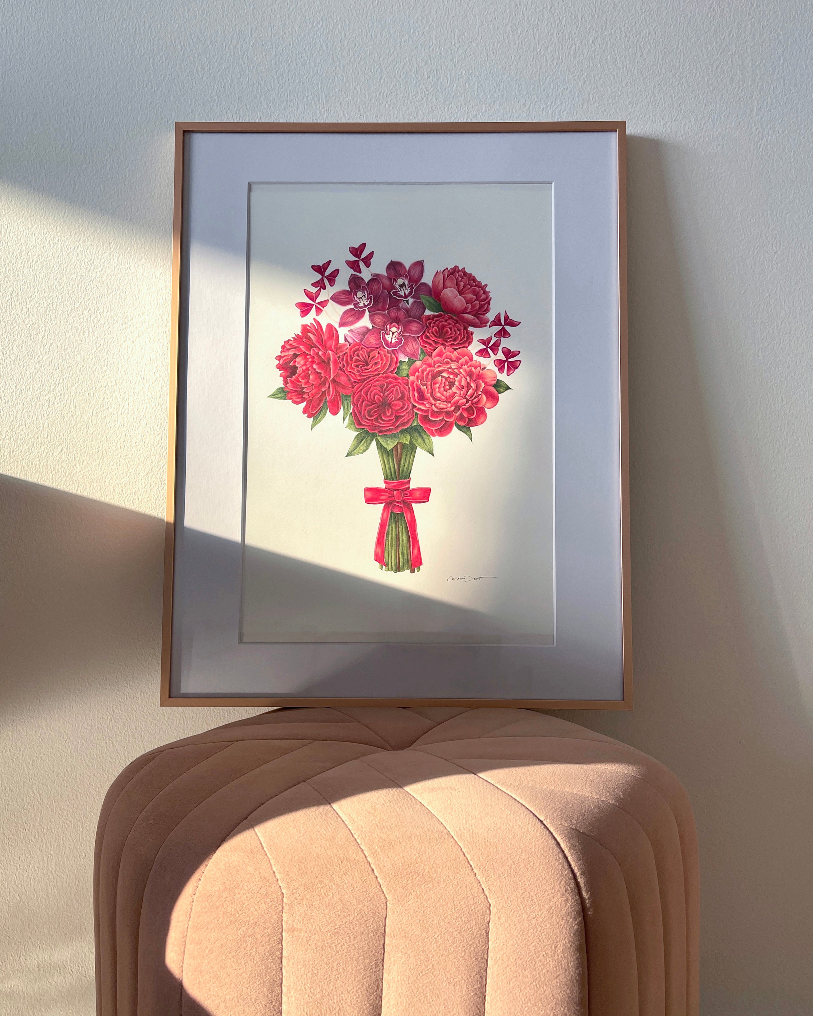 A framed bridal bouquet drawing featuring red flowers tied with a red ribbon, displayed on a pink upholstered stool against a light wall with natural sunlight and shadows.