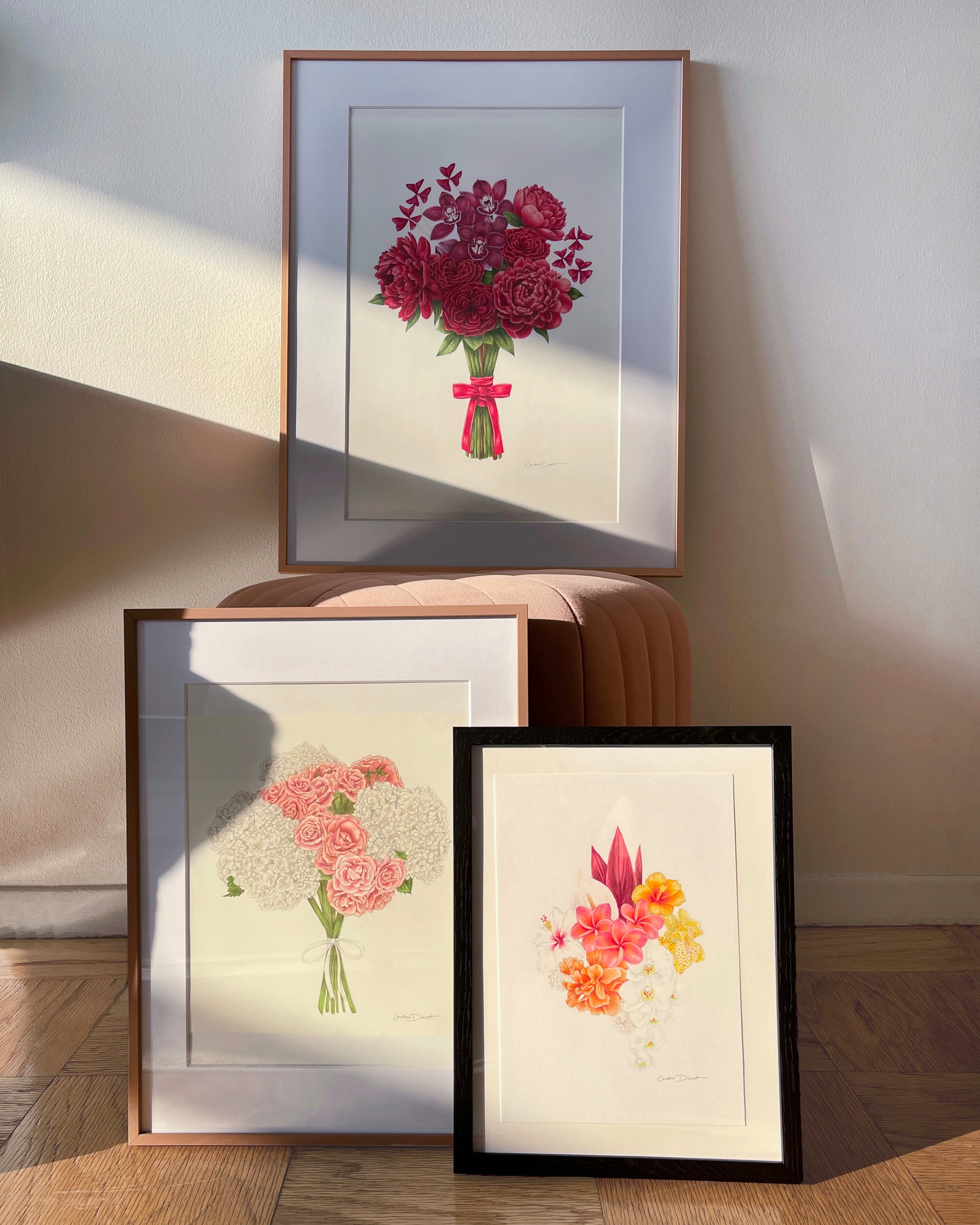 Three framed bridal bouquet drawings are displayed on a wooden floor against a light wall, with one print propped on a small upholstered stool. 