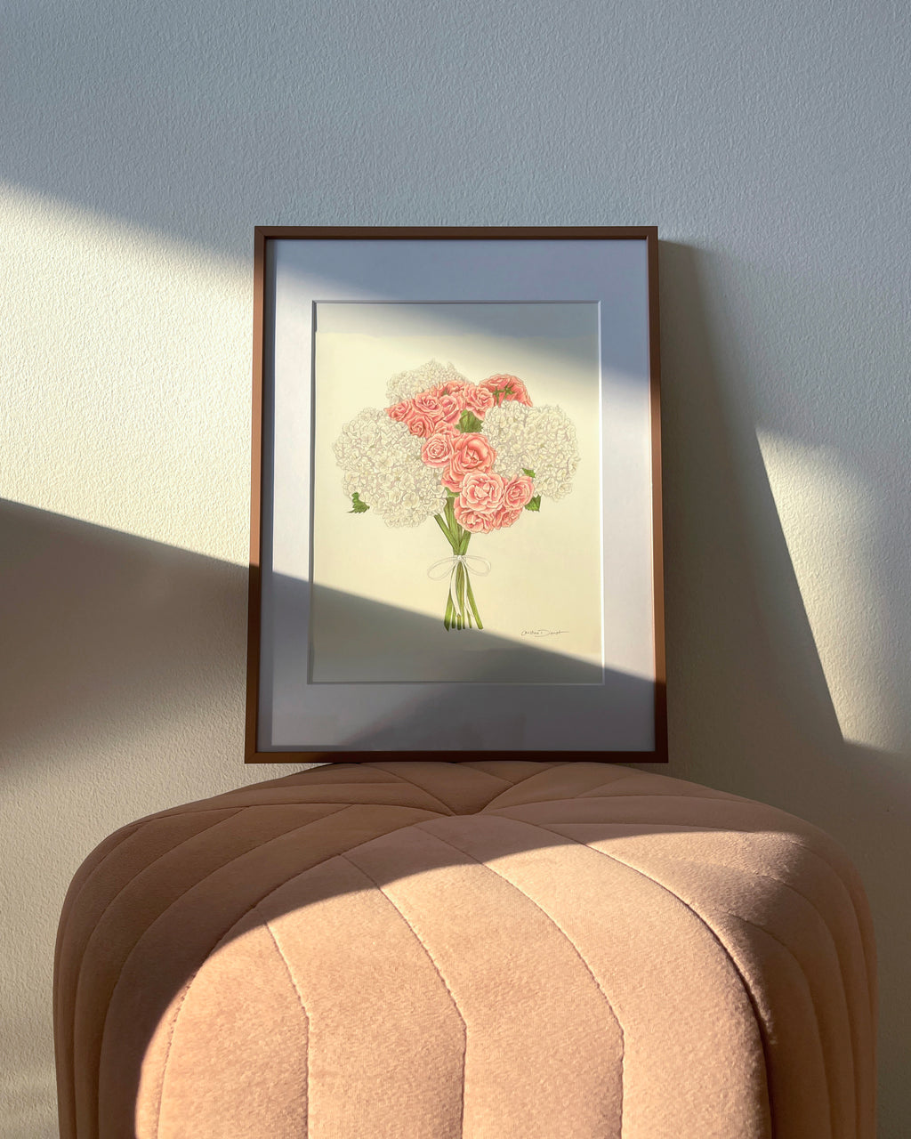 A framed bridal bouquet colored pencil drawing of pink roses and white hydrangeas, displayed on a blush velvet ottoman against a textured wall with soft sunlight and shadows.