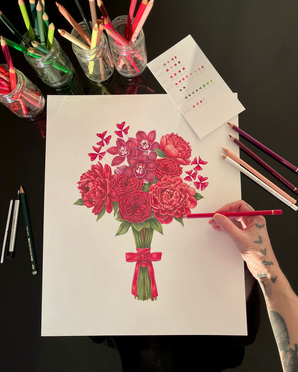 Artist coloring a bouquet of red flowers with colored pencils on a dark surface.
