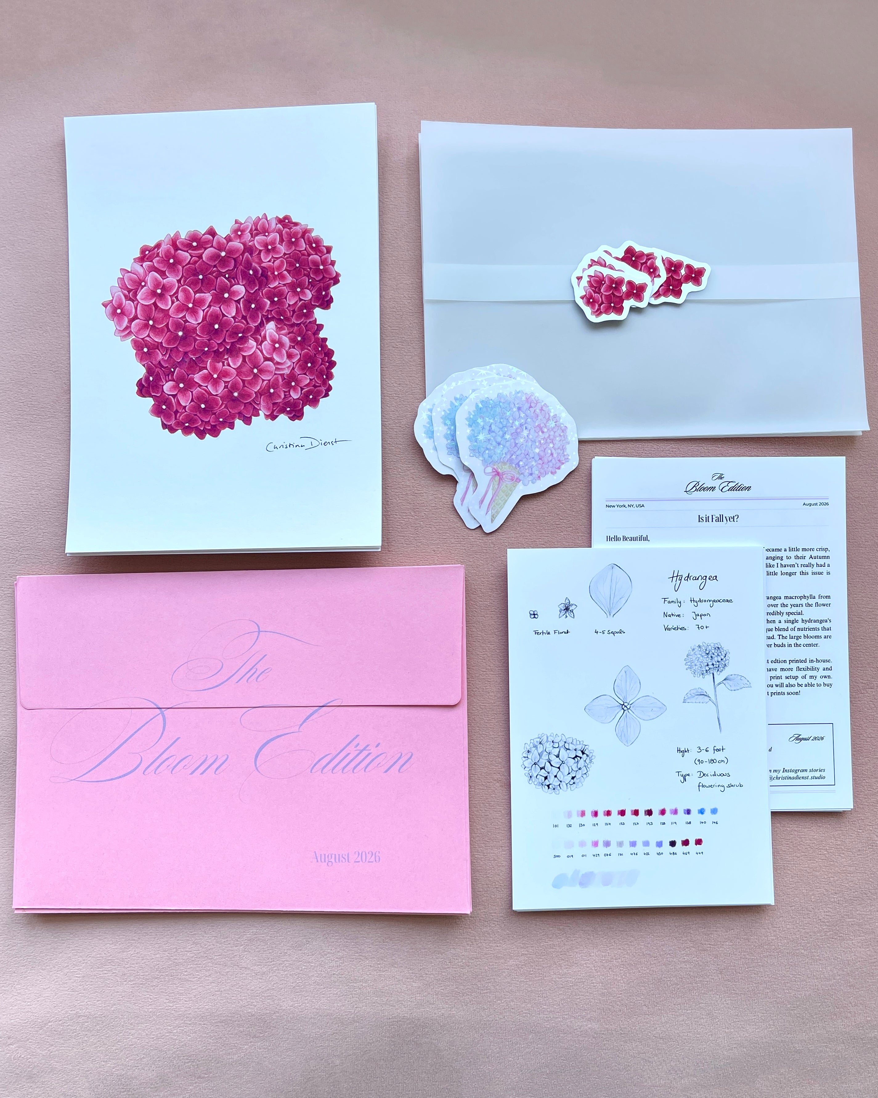 'The Bloom Edition August 2026' arranged on a blush background. The edition includes a pink envelope with blue script, a vellum sleeve with floral sticker seal, a pink hydrangea art print, a pastel flower-shaped sticker, and printed studio note