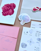 'The Bloom Edition August 2026' arranged on a blush background. The edition includes a pink envelope with blue script, a vellum sleeve with floral sticker seal, a pink hydrangea art print, a pastel flower-shaped sticker, and printed studio note