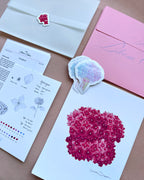 'The Bloom Edition August 2026' arranged on a blush background. The edition includes a pink envelope with blue script, a vellum sleeve with floral sticker seal, a pink hydrangea art print, a pastel flower-shaped sticker, and printed studio note
