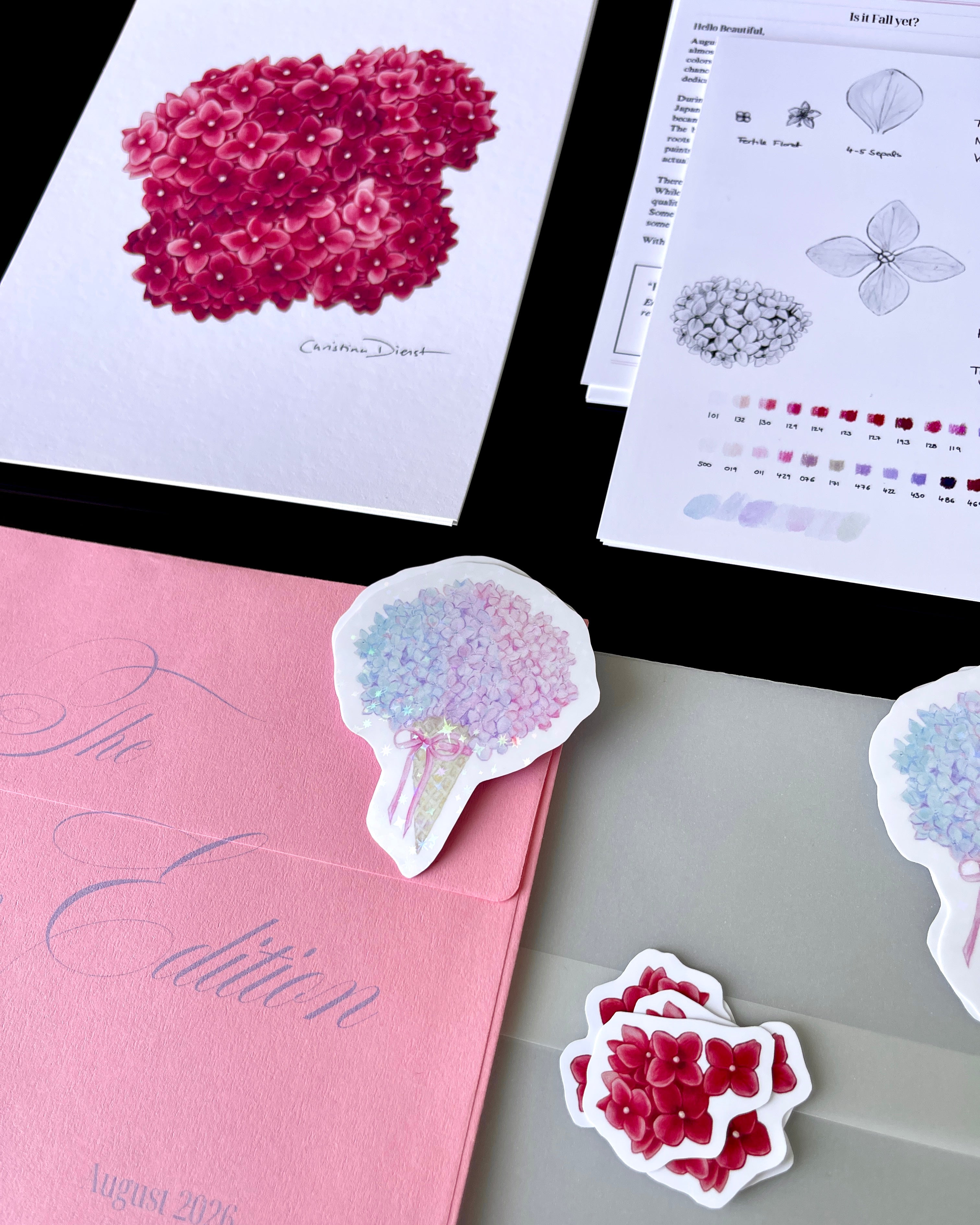 'The Bloom Edition August 2026' arranged on a black background. The edition includes a pink envelope with blue script, a vellum sleeve with floral sticker seal, a pink hydrangea art print, a pastel flower-shaped sticker, and printed studio note