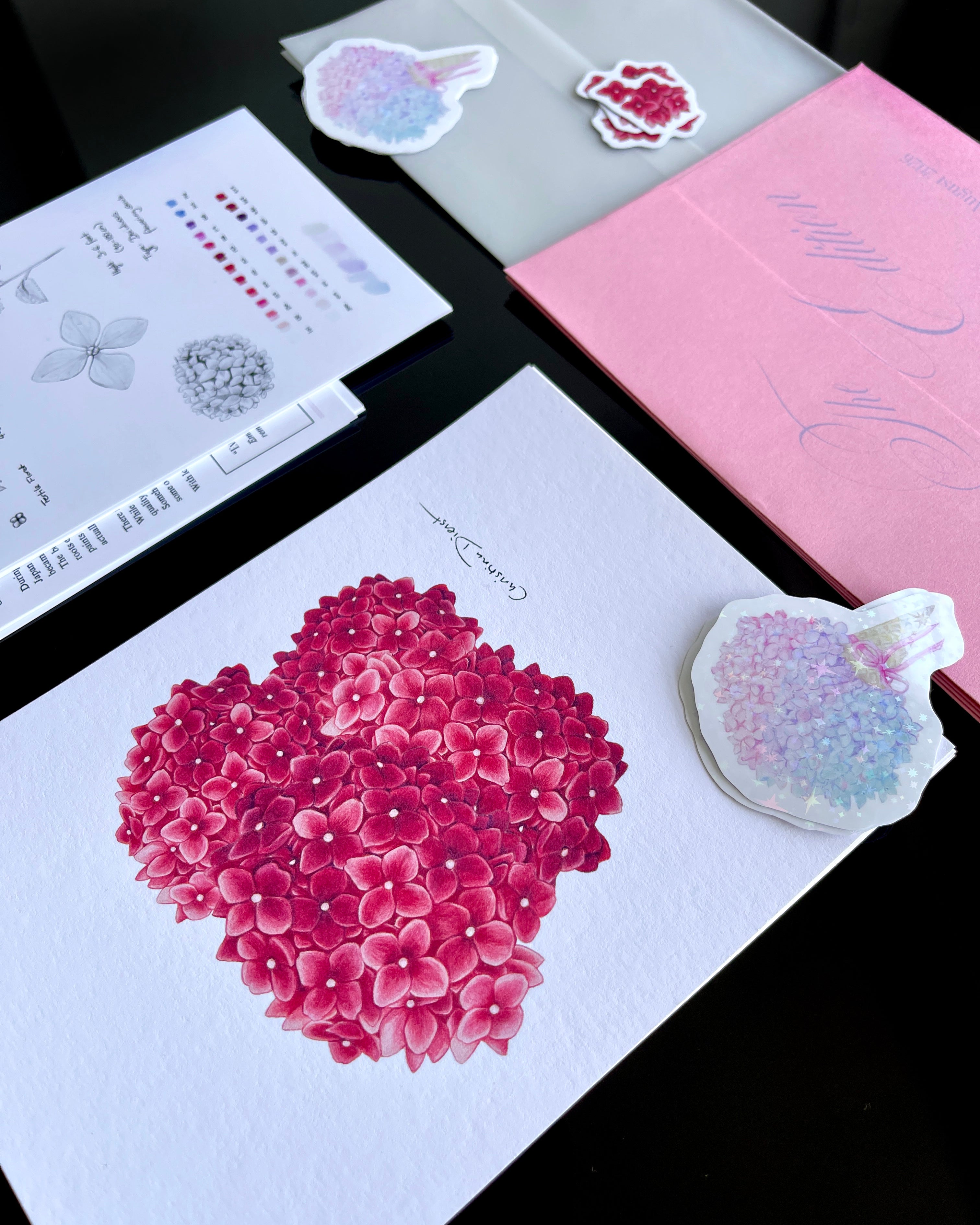 'The Bloom Edition August 2026' arranged on a black background. The edition includes a pink envelope with blue script, a vellum sleeve with floral sticker seal, a pink hydrangea art print, a pastel flower-shaped sticker, and printed studio note
