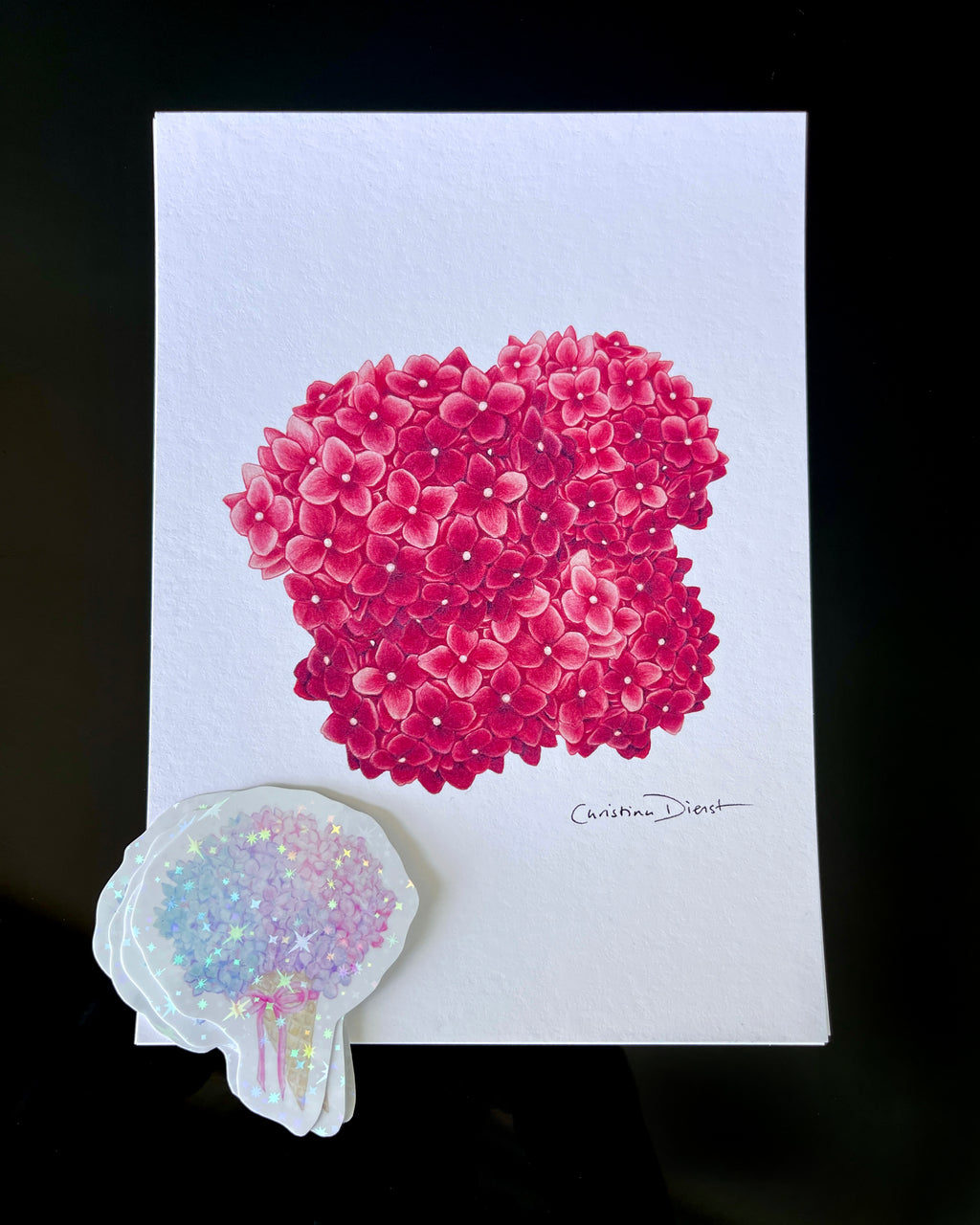 'The Bloom Edition August 2026' arranged on a black background. The edition includes a pink hydrangea art print and a pastel flower-shaped sticker